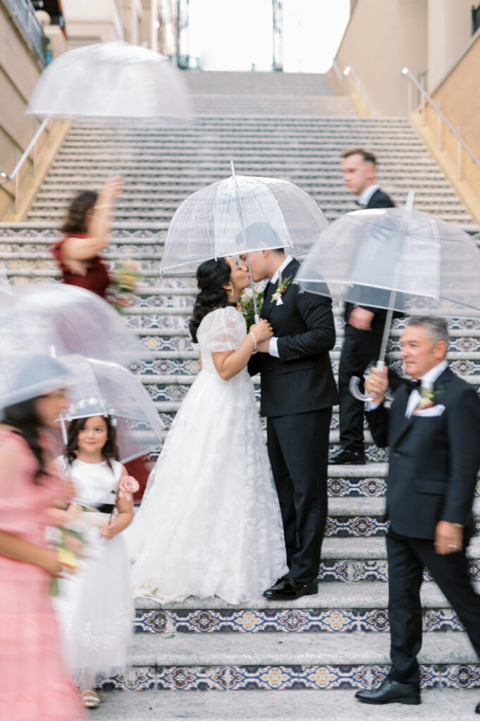 slowmotion image of a wedding party crossing in front and behind the wedding couple while they're kissing on the stairs in the country club plaza in kansas city